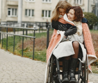 Kobieta przytula inną kobietę, siedzącą na wózku inwalidzkim. W tle trawnik i chodnik prowadzący do bloku mieszkalnego.